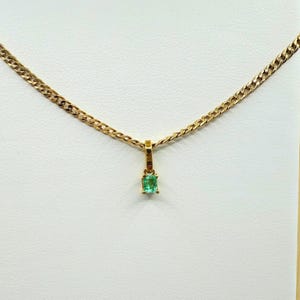 May include: A gold-colored necklace with a small, square emerald pendant. The chain has a curb link design. The pendant is a vibrant green color and is set in a gold-colored setting. The necklace is displayed against a white background.