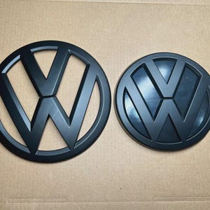 Puede incluir: Dos emblemas negros de Volkswagen, uno calado y otro macizo, sobre un fondo neutro. Ambos presentan el logotipo de VW. Ideales para entusiastas del automóvil y coleccionistas.