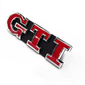 May include: A GTI emblem with a black base and the letters GTI in red with a chrome border. The emblem is angled on a white background. The letters are bold and glossy.