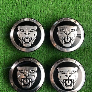 Puede incluir: Cuatro tapacubos de coche Jaguar. Cada tapacubo presenta un emblema de cabeza de jaguar plateado sobre un fondo negro, rodeado por un anillo plateado con la palabra "JAGUAR". Los tapacubos están colocados sobre una superficie verde.