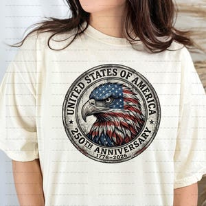 Op de afbeelding: Crèmekleurig T-shirt met een cirkelvormig embleem met een adelaarskop. Het embleem bevat de tekst "UNITED STATES OF AMERICA" en "250TH ANNIVERSARY 1776-2026". De kop van de adelaar is gekleurd in de Amerikaanse vlag.