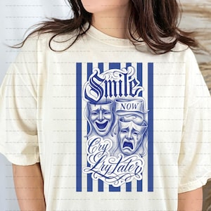 Könnte beinhalten: Cremefarbenes T-Shirt mit blauem Aufdruck. Der Aufdruck zeigt die Worte "Smile Now, Cry Later" über Bildern eines lachenden und eines weinenden Theatermaske. Vertikale blaue Streifen umrahmen das Design.