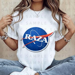 May include: White t-shirt featuring a blue and orange graphic with the word "RAZA" in white. The design is a parody of the NASA logo. The shirt is made of a soft material and has short sleeves.