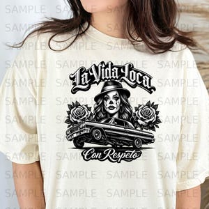 Peut inclure: T-shirt crème avec un motif noir et blanc. Le motif comprend le texte "La Vida Loca" au-dessus d'une image d'une femme portant un chapeau, une voiture lowrider et les mots "Con Respeto". Deux roses encadrent l'image centrale.