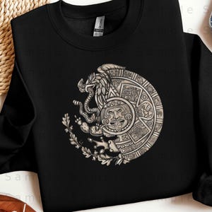 May include: Black crewneck sweatshirt featuring a detailed, circular graphic. The graphic depicts a stylized eagle with a snake, a face, and intricate patterns, likely representing cultural or historical symbolism. The design is in shades of gray and white.