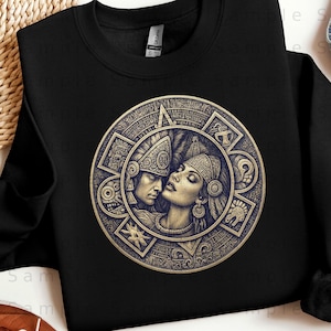May include: Black crewneck sweatshirt featuring a detailed, circular design with an Aztec-inspired illustration. The design depicts two figures in profile, surrounded by intricate patterns and symbols in shades of gold and brown.