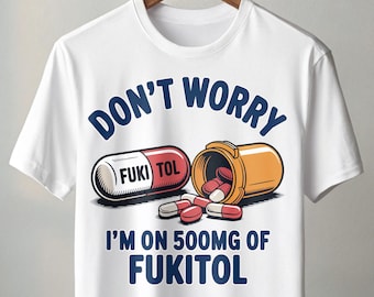 May include: White t-shirt with a humorous graphic. The design features a large capsule with "FUKI TOL" printed on it, a pill bottle, and the text "DON'T WORRY I'M ON 500MG OF FUKITOL" in blue. The shirt is hanging on a wooden hanger.