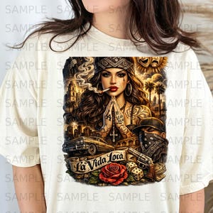 May include: Cream-colored t-shirt featuring a detailed graphic. The design includes a woman with tattoos, a bandana, and a cigarette, with the text "La Vida Loca" on a banner, a rose, a lowrider car, and a gun.