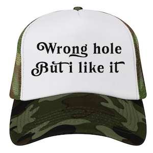 Op de afbeelding: Truckerpet met een wit voorpaneel en een camouflageklep en mesh achterkant. De pet heeft de tekst "Wrong hole But I like it" in zwart.