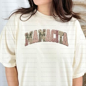 Könnte beinhalten: Cremefarbenes T-Shirt mit dem Wort "MAMACITA" in einem Bogen. Der Text ist in einer Schriftart im College-Stil mit einem Tarn- und Karomuster gehalten. Das Shirt hat kurze Ärmel.