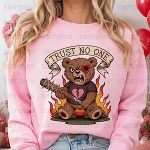 Puede incluir: Sudadera rosa con un gráfico de oso de peluche marrón. El oso tiene una expresión de enojo, un parche en el ojo en forma de corazón y un corazón roto en su camisa. Sostiene un bate con púas, con llamas alrededor. El texto "TRUST NO ONE" está encima del oso.