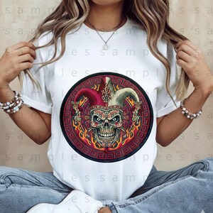 May include: White t-shirt featuring a circular graphic of a skull wearing a jester's hat. The skull is detailed with intricate designs and surrounded by flames and a decorative border. The design incorporates red, burgundy, and gold colors.