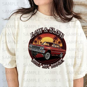 以下が含まれることがあります： クリーム色のTシャツ。赤いクラシックカーのグラフィックと「LOW & SLOW」と「BUILT NOT BOUGHT」の文字が円形にデザインされています。デザインにはヤシの木と夕焼けが含まれています。