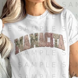 May include: White t-shirt with the word "MAMACITA" in a collegiate style font. The letters are filled with a mix of patterns, including camouflage and plaid, and outlined in a darker color.