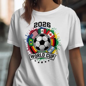 May include: White t-shirt featuring a soccer ball design with the text "2026 WORLD CUP." The design includes flags of various countries and colorful paint splatters. The shirt is a crew neck style.