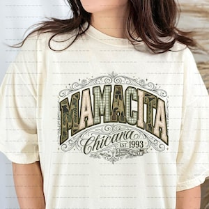 Puede incluir: Camiseta color crema con el texto "MAMACITA Chicana EST. 1993 BARRIO STYLE" en un diseño decorativo. La palabra "MAMACITA" tiene un patrón de camuflaje. La camiseta tiene mangas cortas y cuello redondo.