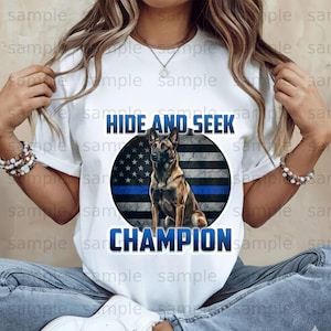 May include: White t-shirt with a dog graphic and the text "HIDE AND SEEK CHAMPION". The design incorporates an American flag with a blue line. The dog is depicted in shades of brown and black.
