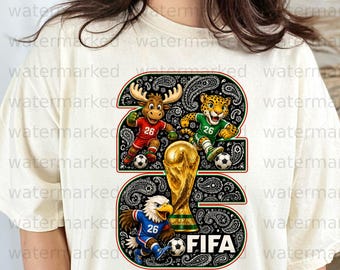 Puede incluir: Camiseta color crema con un gran gráfico del número 26 y mascotas de animales jugando al fútbol. El diseño incluye un alce, un jaguar y un águila, junto con un trofeo dorado y la palabra "FIFA".