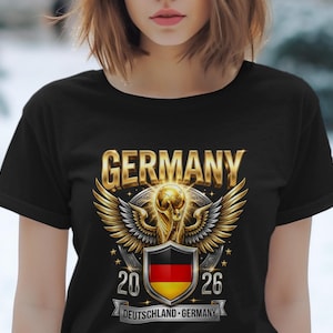 Peut inclure: T-shirt noir avec un motif doré et argenté. Le mot "GERMANY" est au-dessus d'un trophée ailé et d'un bouclier avec le drapeau allemand. Le t-shirt affiche également les années "20" et "26" et le texte "DEUTSCHLAND-GERMANY".