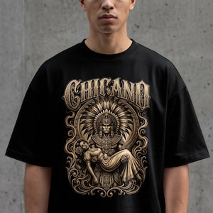 Puede incluir: Una camiseta negra con un estampado gráfico en tonos dorados. El diseño muestra la palabra CHICANO en una fuente estilizada sobre la ilustración de una figura con un tocado de plumas que sostiene a otra figura, rodeada de adornos en espiral.
