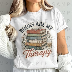 May include: White t-shirt with a graphic of a book stack, flowers, and a coffee cup. The text "BOOKS ARE MY Therapy" is printed in a circular design. The shirt is a casual style.
