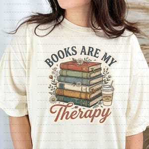 May include: Cream-coloured t-shirt with a stack of books and the words "Books Are My Therapy". A coffee cup with "Bookshop Cafe" is next to the books. Floral accents surround the books, all in a vintage style.