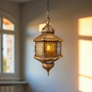 May include: Ornate, gold-colored pendant lamp with intricate cut-out patterns. The lamp hangs from a chain and illuminates with a warm, inviting glow. The lamp's design is reminiscent of Moroccan or Middle Eastern styles, adding a touch of exotic elegance to the room.