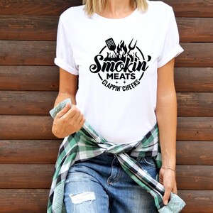 May include: White t-shirt with a black graphic design. The design features a circular emblem with a spatula, fork, and flames, and the words "Smokin' Meats Clappin' Cheeks". The shirt is paired with blue jeans and a green and black plaid shirt tied around the waist.
