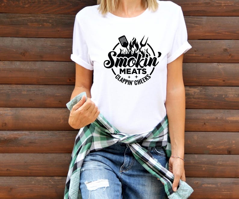 May include: White t-shirt with a black graphic design. The design features a circular emblem with a spatula, fork, and flames, and the words "Smokin' Meats Clappin' Cheeks". The shirt is paired with blue jeans and a green and black plaid shirt tied around the waist.