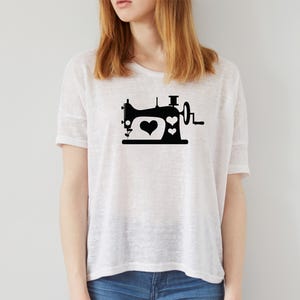 Op de afbeelding: Wit T-shirt met een zwarte afbeelding van een vintage naaimachine. Het ontwerp van de naaimachine bevat twee harten. Het shirt is gemaakt van licht materiaal en heeft een losse pasvorm. Het model draagt een blauwe jeans.