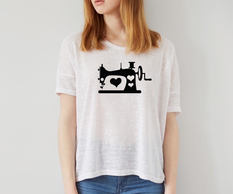 Op de afbeelding: Wit T-shirt met een zwarte afbeelding van een vintage naaimachine. Het ontwerp van de naaimachine bevat twee harten. Het shirt is gemaakt van licht materiaal en heeft een losse pasvorm. Het model draagt een blauwe jeans.