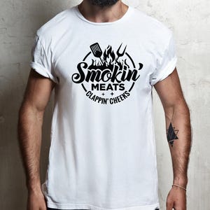 May include: White t-shirt with a black graphic design. The design features the words "Smokin' Meats, Clappin' Cheeks" in a circular arrangement, with a spatula, fork, and flames above the text. The shirt has rolled-up sleeves.