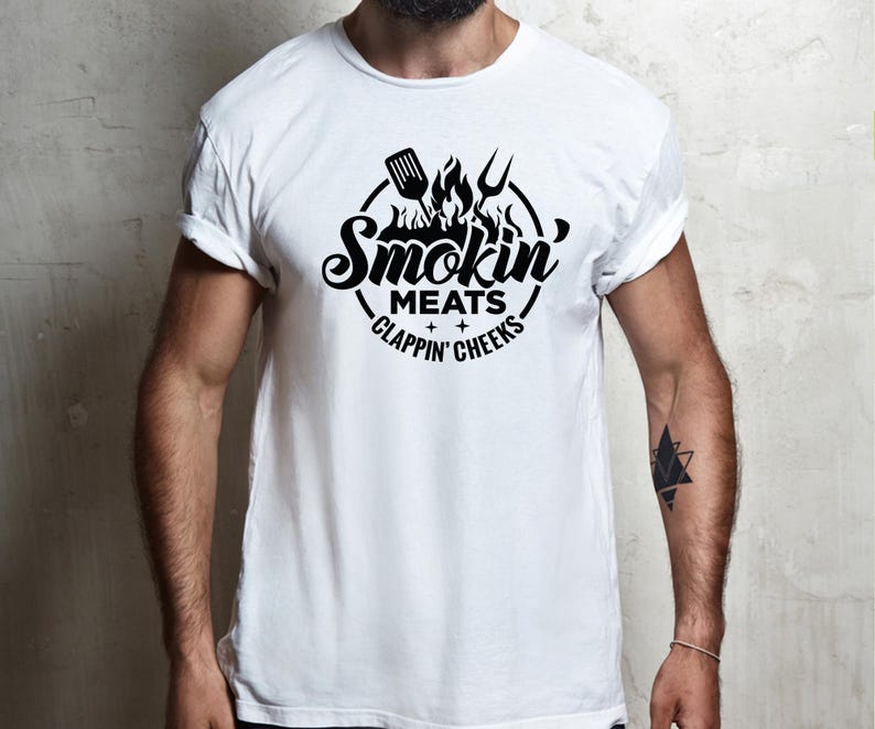 May include: White t-shirt with a black graphic design. The design features the words "Smokin' Meats, Clappin' Cheeks" in a circular arrangement, with a spatula, fork, and flames above the text. The shirt has rolled-up sleeves.