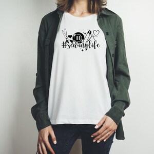 Op de afbeelding: Wit T-shirt met een zwarte afbeelding van naai-attributen, een knoop en de tekst "#sewinglife". Het ontwerp bevat een schaar, een hart en een naald met draad. De persoon draagt een groene jas en een donkerblauwe jeans.