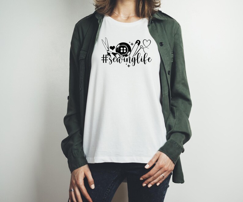 Op de afbeelding: Wit T-shirt met een zwarte afbeelding van naai-attributen, een knoop en de tekst "#sewinglife". Het ontwerp bevat een schaar, een hart en een naald met draad. De persoon draagt een groene jas en een donkerblauwe jeans.