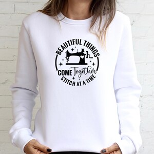 Op de afbeelding: Witte sweatshirt met een zwarte cirkelvormige afbeelding met een naaimachine, een hartslag en de tekst "BEAUTIFUL THINGS COME TOGETHER STITCH AT A TIME."