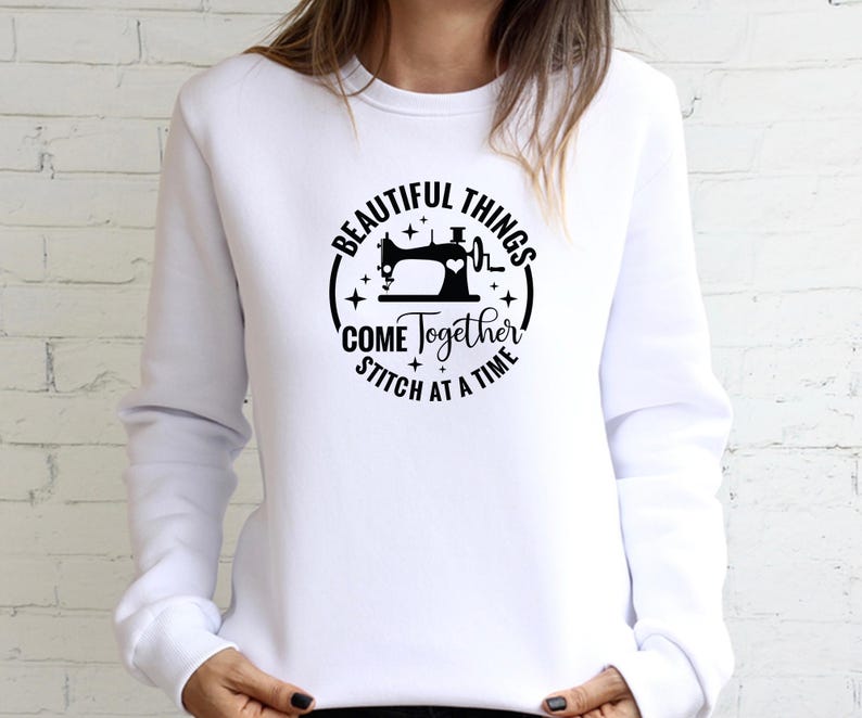 Op de afbeelding: Witte sweatshirt met een zwarte cirkelvormige afbeelding met een naaimachine, een hartslag en de tekst "BEAUTIFUL THINGS COME TOGETHER STITCH AT A TIME."