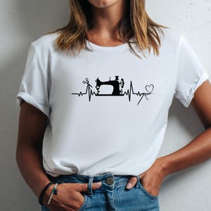 Op de afbeelding: Wit T-shirt met een zwarte afbeelding van een naaimachine, schaar en een hart, ge&iuml;ntegreerd in een hartslaglijnontwerp. Het shirt is gemaakt van een zacht materiaal en heeft korte mouwen.