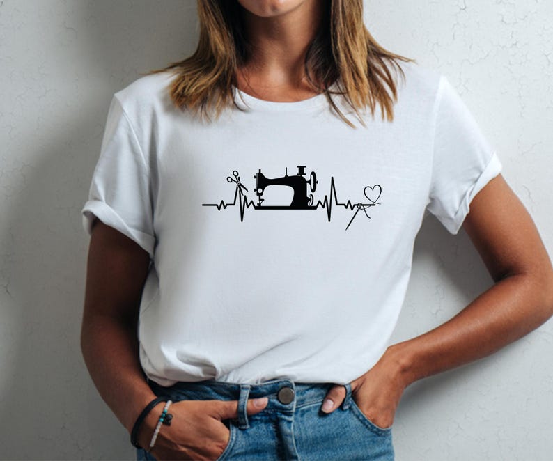 Op de afbeelding: Wit T-shirt met een zwarte afbeelding van een naaimachine, schaar en een hart, ge&iuml;ntegreerd in een hartslaglijnontwerp. Het shirt is gemaakt van een zacht materiaal en heeft korte mouwen.