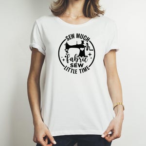 Op de afbeelding: Wit t-shirt met een zwart grafisch ontwerp met een naaimachine, een hart en de woorden "Sew Much Fabric, Sew Little Time" in een cirkelvormig ontwerp. Het shirt heeft korte mouwen en een ronde hals.