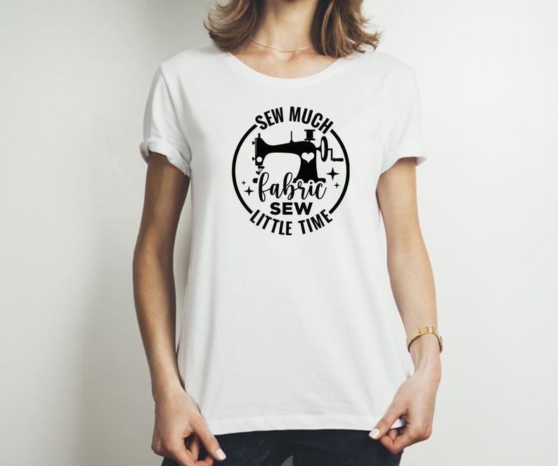 Op de afbeelding: Wit t-shirt met een zwart grafisch ontwerp met een naaimachine, een hart en de woorden "Sew Much Fabric, Sew Little Time" in een cirkelvormig ontwerp. Het shirt heeft korte mouwen en een ronde hals.