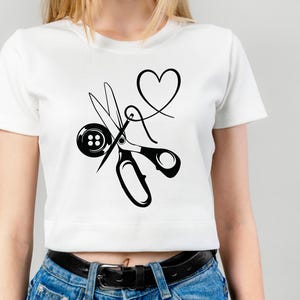 Op de afbeelding: Wit T-shirt met een zwarte afbeelding van naai-attributen. Het ontwerp bevat een schaar, een knoop, een naald en draad in de vorm van een hart. Het shirt is een cropped model.