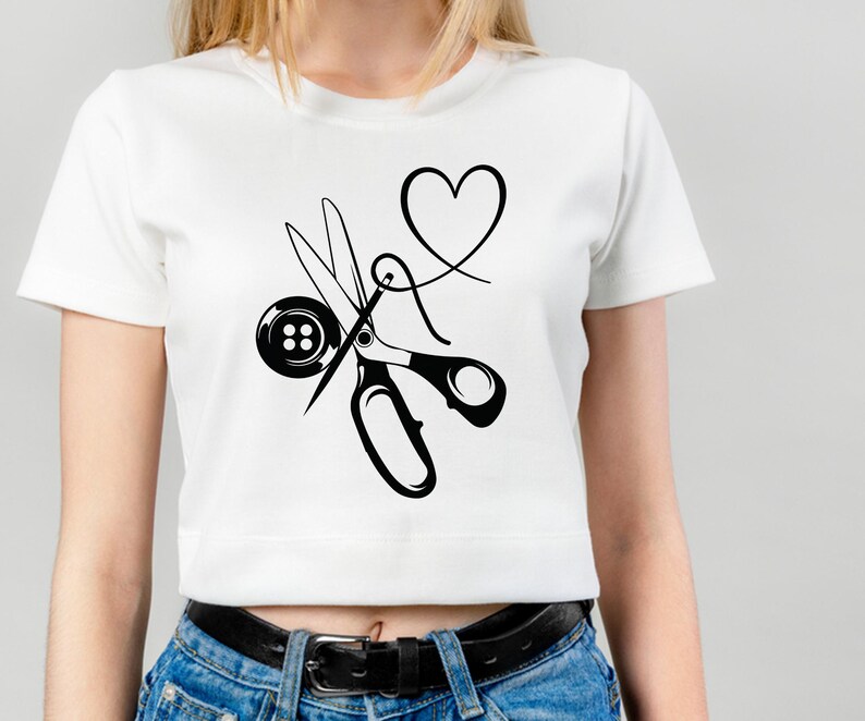 Op de afbeelding: Wit T-shirt met een zwarte afbeelding van naai-attributen. Het ontwerp bevat een schaar, een knoop, een naald en draad in de vorm van een hart. Het shirt is een cropped model.