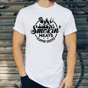 May include: White t-shirt with a black graphic design. The design features a circular emblem with the words "Smokin' Meats Clappin' Cheeks" and images of a spatula, fork, and flames. The shirt is short-sleeved.