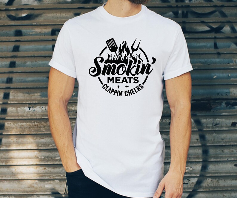 May include: White t-shirt with a black graphic design. The design features a circular emblem with the words "Smokin' Meats Clappin' Cheeks" and images of a spatula, fork, and flames. The shirt is short-sleeved.