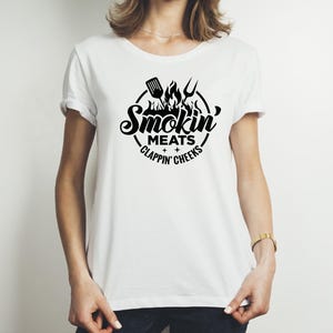 May include: White t-shirt with a black graphic design. The design features the words "Smokin' Meats + Clappin' Cheeks" in a circular arrangement, with a spatula, flames, and a fork. The shirt is a casual, crew neck style.
