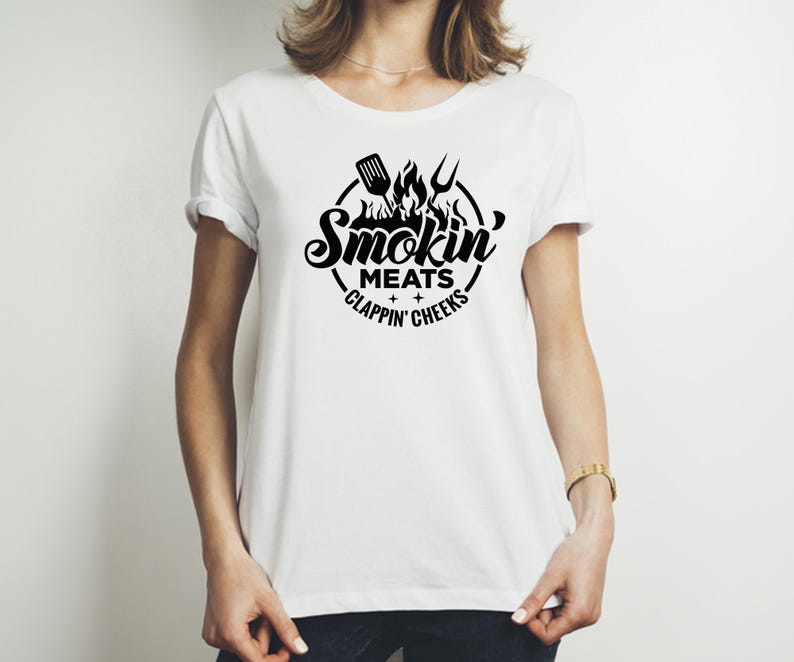 May include: White t-shirt with a black graphic design. The design features the words "Smokin' Meats + Clappin' Cheeks" in a circular arrangement, with a spatula, flames, and a fork. The shirt is a casual, crew neck style.