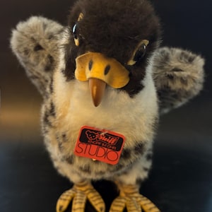 Puede incluir: Un juguete de peluche de pájaro con una cabeza marrón oscuro, alas grises y blancas y un pecho color crema. Tiene un pico y patas amarillas. Una etiqueta roja con la palabra "STUDIO" está adjunta. El juguete está diseñado para parecerse a un halcón.