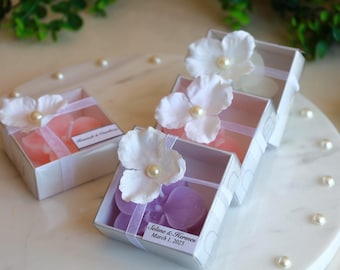 May include: White boxes with clear lids, each containing a soap or candy, are decorated with a white flower and a pearl. The boxes are tied with a sheer ribbon and sit on a white marble surface.