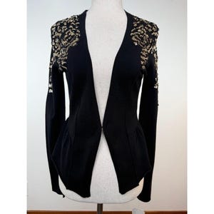 May include: A black long-sleeved cardigan featuring a deep V-neckline and a single hook-and-eye closure at the center. The shoulders are adorned with intricate gold sequin embellishments that extend down the upper sleeves in a floral pattern.
